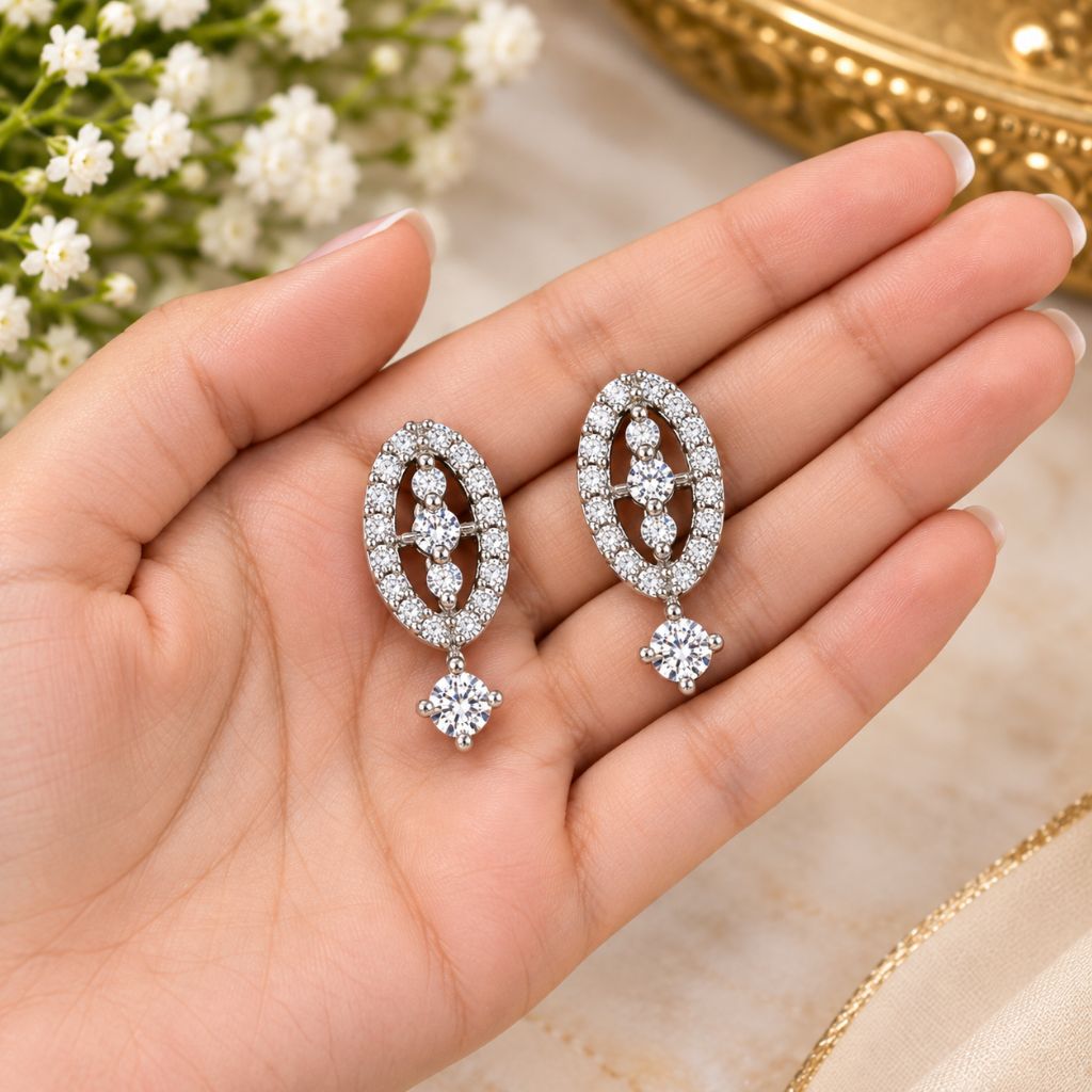 A pair of silver-tone oval drop earrings paved with clear stones, held in a person's hand against a soft beige background with small white flowers and a gold decorative object in the background.