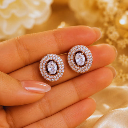 Rose gold earrings with clear gemstones held between fingers against a blurred gold background
