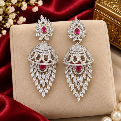 Affordable diamond earrings with ruby pink stones and American Diamond jhumka design. A pair of ornate silver-toned dangle earrings with red gemstones and clear stones, displayed on a beige jewelry box with red satin fabric and pearl-like beads around them.