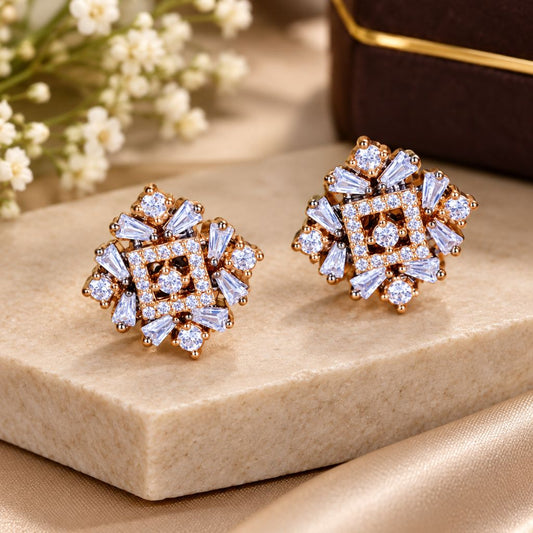 A pair of gold-toned geometric stud earrings with clear baguette and round stones, displayed on a beige stone block with soft satin fabric and small white flowers in the background.