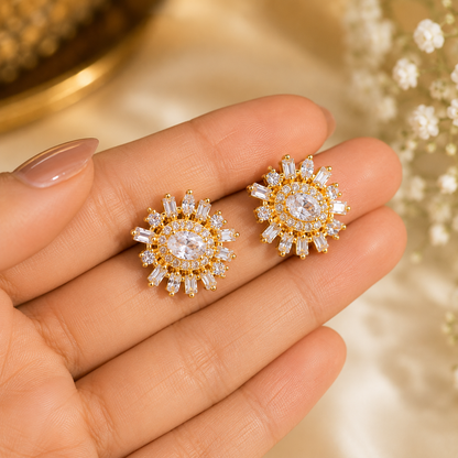 Gold-plated American diamond stud earrings with round-cut sparkling stones and baguette accents displayed on a hand.