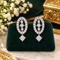 A pair of silver-toned oval drop earrings paved with clear stones and featuring a dangling round-cut stone, displayed on a dark green velvet stand with white flowers and gold decor in the background.