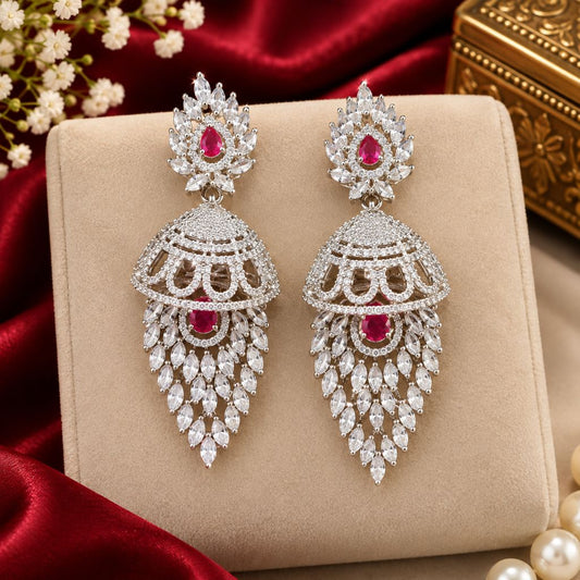 A pair of ornate silver-toned dangle earrings with red gemstones and clear stones, displayed on a beige jewelry box with red satin fabric and pearl-like beads around them.