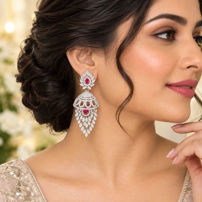 A close-up lifestyle shot of a woman wearing a large, ornate silver-toned jhumka earring adorned with clear stones and a central pinkish-red stone, set against a softly blurred background.