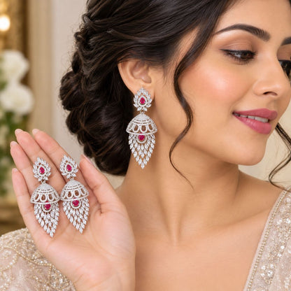 A woman wearing a matching pair of silver-toned chandelier earrings with red gemstone accents, holding the earrings in her hand against a softly blurred indoor background.