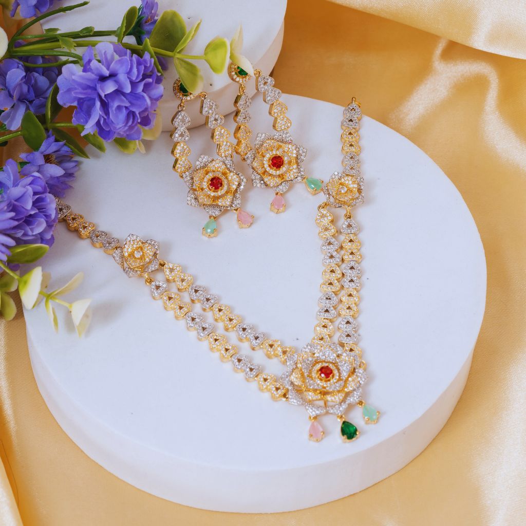 Gold and silver beaded jewelry set with red and green gemstone floral motifs, displayed on a white round pedestal with purple flowers and yellow satin fabric.
