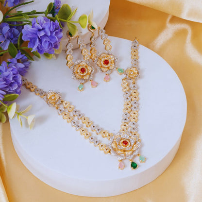 Gold and silver beaded jewelry set with red and green gemstone floral motifs, displayed on a white round pedestal with purple flowers and yellow satin fabric.