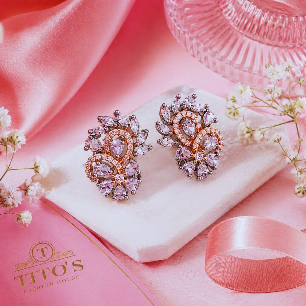 A pair of ornate floral stud earrings with pink and clear rhinestones on a white display cushion, styled on a pink satin surface with small white flowers, a pink ribbon, and a pink glass dish. The brand name 'TITO'S FASHION HOUSE' is visible on a pink card in the lower left.