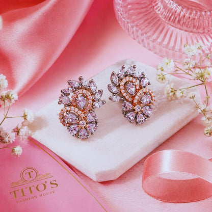 A pair of ornate floral stud earrings with pink and clear rhinestones on a white display cushion, styled on a pink satin surface with small white flowers, a pink ribbon, and a pink glass dish. The brand name 'TITO'S FASHION HOUSE' is visible on a pink card in the lower left.
