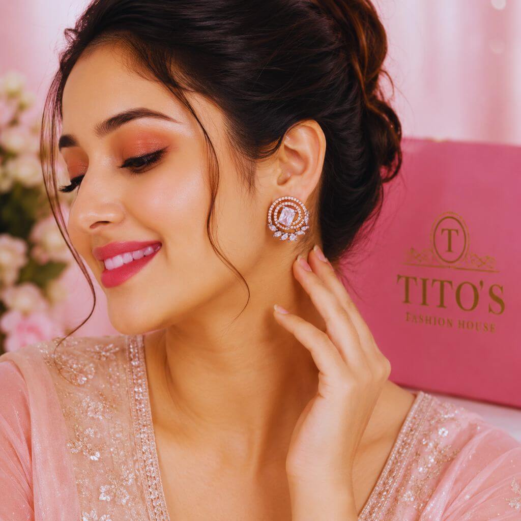 A woman in a pink embellished top wears a circular rose-gold-toned earring with clear stones. A pink shopping bag with the brand name 'TITO'S FASHION HOUSE' is visible in the background.