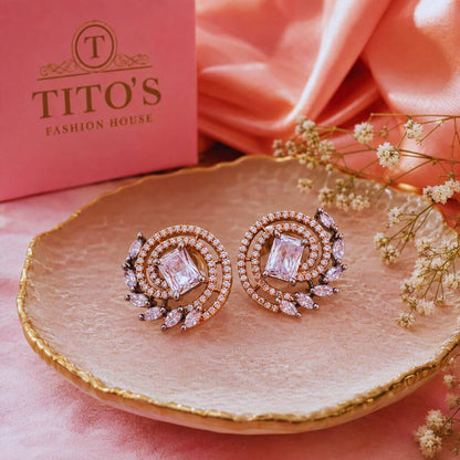 A pair of rose gold crescent-shaped stud earrings with large emerald-cut pink stones and surrounding clear crystals, displayed on a gold-rimmed pink plate. A pink box labeled 'TITO'S FASHION HOUSE' is visible in the background along with peach fabric and small white flowers.