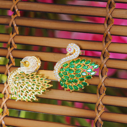 Gold and green peacock-shaped earrings on a woven surface with colorful background