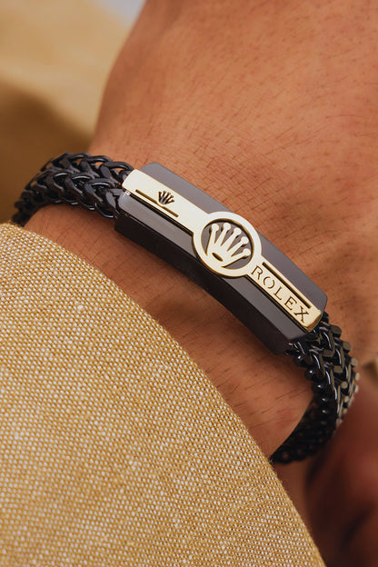 Black braided bracelet with gold tag featuring Rolex logo on wrist against beige fabric background