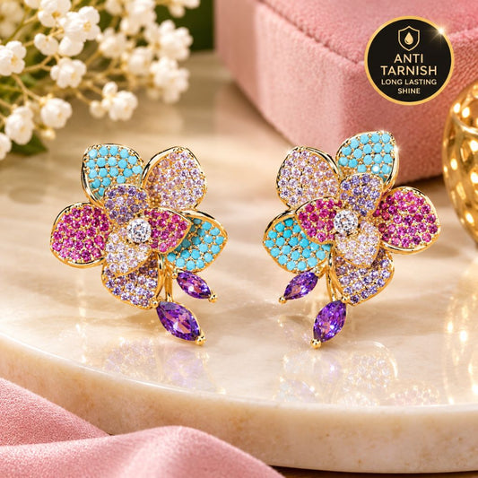 A pair of gold-tone floral stud earrings decorated with pink, turquoise, and lavender pavé stones, displayed on a polished cream marble slab with pink fabric and white flowers in the background. A circular badge in the top right reads 'ANTI TARNISH LONG LASTING SHINE'.