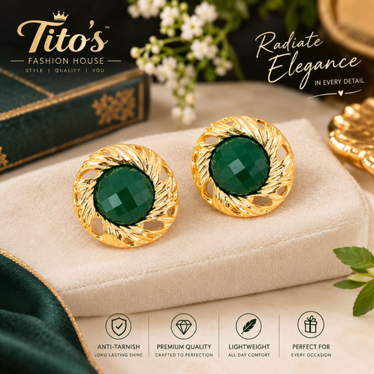 A pair of gold-toned oval stud earrings with faceted dark green centers, displayed on a beige fabric surface with decorative props and branding text for Tito's Fashion House.