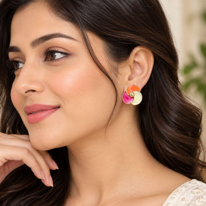 A close-up of a woman wearing a pair of colorful, layered circular stud earrings in orange, pink, and cream, with her hand resting near her chin.