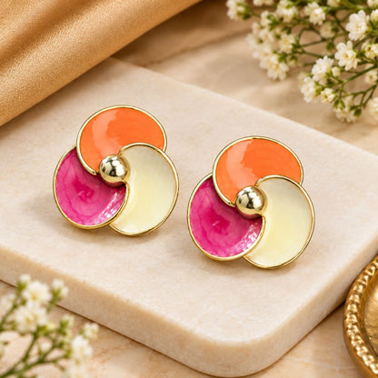 A pair of gold-tone circular stud earrings with overlapping enamel sections in orange, pink, and cream, displayed on a beige stone slab with small white flowers and a gold fabric accent.