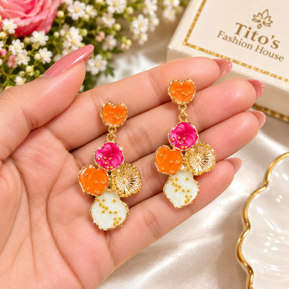 A pair of gold-tone drop earrings with colorful enamel flower and fruit charms, held in a hand. The earrings feature orange and pink flowers, gold leaf details, and a white enamel petal with gold speckles.