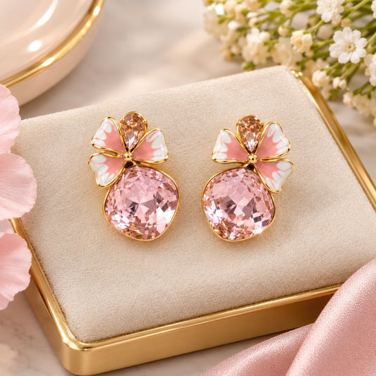 A pair of gold-tone stud earrings with large faceted pink gemstones and bow-shaped pink, white, and gold enamel tops, displayed on a cream jewelry cushion in a gold box with soft pink flowers around them.