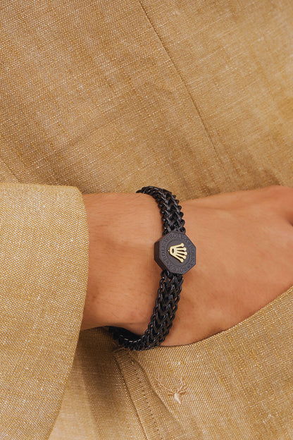 Black braided bracelet with a hexagonal emblem on a wrist against a beige fabric background