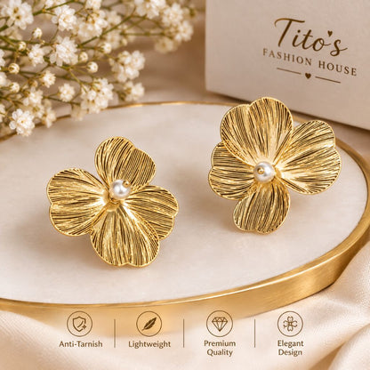 A pair of gold flower-shaped stud earrings with a small white pearl at the center, displayed on a gold-rimmed cream tray. The background features a beige box for 'Tito's Fashion House' and delicate white baby's breath flowers.