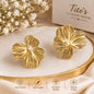 A pair of gold flower-shaped stud earrings with a small white pearl at the center, displayed on a gold-rimmed cream tray. The background features a beige box for 'Tito's Fashion House' and delicate white baby's breath flowers.