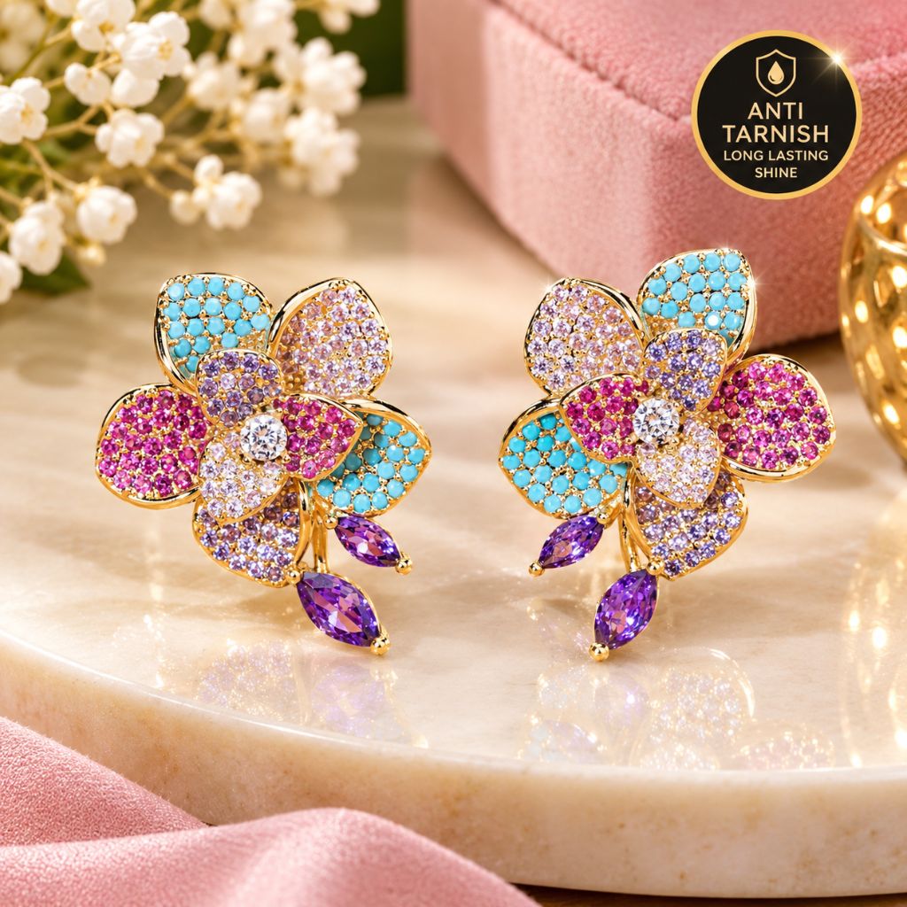 A pair of gold-tone floral stud earrings decorated with pink, turquoise, and lavender pavé stones, displayed on a polished cream marble slab with pink fabric and white flowers in the background. A circular badge in the top right reads 'ANTI TARNISH LONG LASTING SHINE'.
