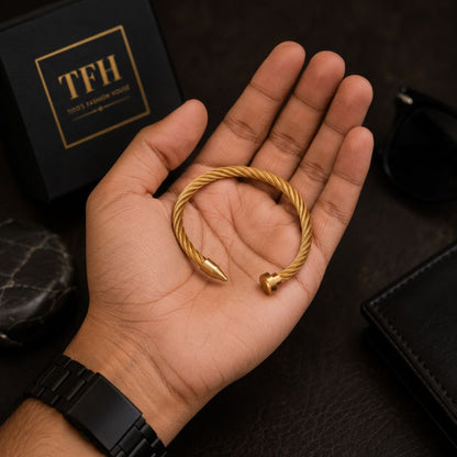 A gold-toned twisted cable open cuff bracelet with arrowhead tips, held in a person's hand. A black TFH box, sunglasses, and a black leather wallet are visible in the background.