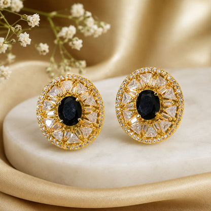 A pair of round gold-toned statement earrings with black oval center stones and clear faceted surrounding stones, displayed on a cream stone slab with soft gold fabric and small white flowers in the background.