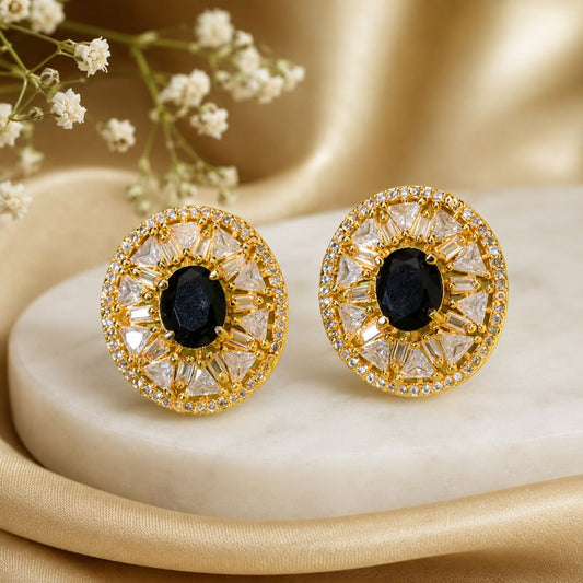 A pair of round gold-toned statement earrings with black oval center stones and clear faceted surrounding stones, displayed on a cream stone slab with soft gold fabric and small white flowers in the background.