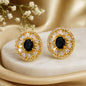 A pair of round gold-toned statement earrings with black oval center stones and clear faceted surrounding stones, displayed on a cream stone slab with soft gold fabric and small white flowers in the background.