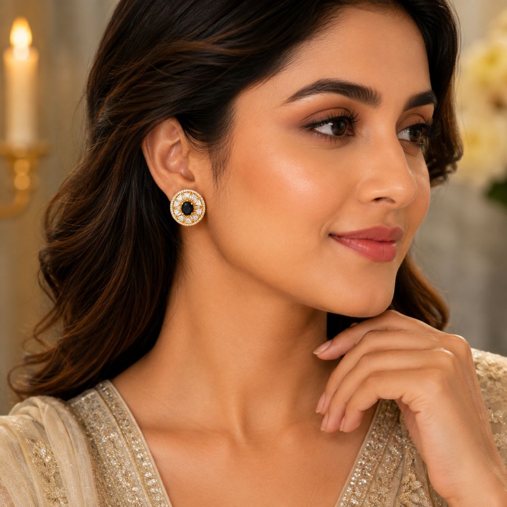 A woman wearing a round gold-toned earring with a black center and small white stones, shown in a close-up portrait against a softly blurred indoor background.