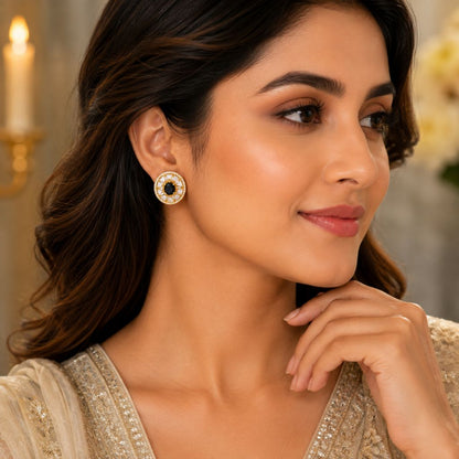 A woman wearing a round gold-toned earring with a black center and small white stones, shown in a close-up portrait against a softly blurred indoor background.