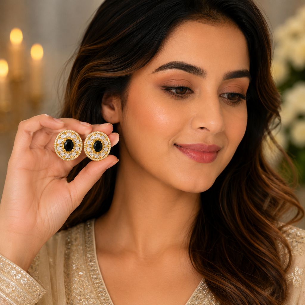 A woman holds a pair of gold-toned statement earrings with black centers and clear stones between her fingers, wearing one earring. She is dressed in a gold embellished top against a softly blurred background with warm lights and flowers.
