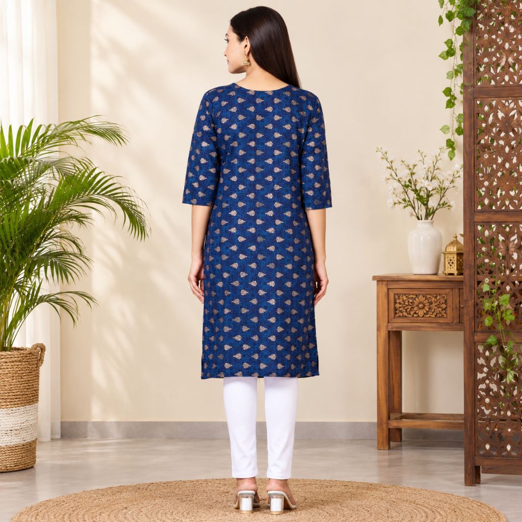 A woman stands with her back to the camera wearing a blue three-quarter sleeve kurta with a gold geometric pattern, paired with white pants, in a styled indoor setting with plants and a wooden screen.