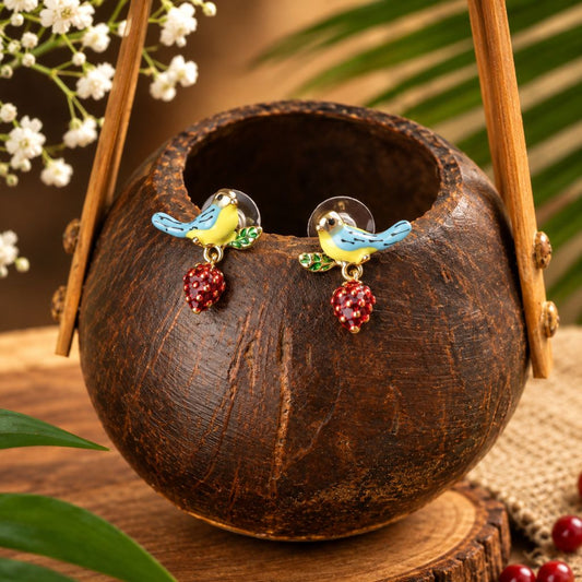 A pair of bird stud earrings displayed on a dark brown coconut bowl. The earrings feature small yellow birds with blue wings and green leaves, each hanging from a red berry charm.