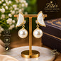 A pair of gold and mother-of-pearl butterfly drop earrings displayed on a gold stand, with a dark box, white flowers, and decorative text overlays in the background.