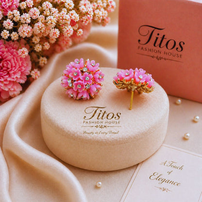 Pink floral earrings on a 'Titos Fashion House' branded box with flowers in the background.