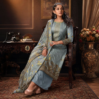 A woman seated in a dark wooden chair wearing a light blue traditional outfit with gold embroidery and a matching dupatta, posed in an ornate room with a vintage desk, framed wall art, and a large floral vase.