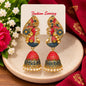 A pair of ornate gold-tone and red-blue jhumka earrings displayed on a white card labeled 'Fashion Earring,' set on a beige dish with flowers and a red patterned fabric in the background.