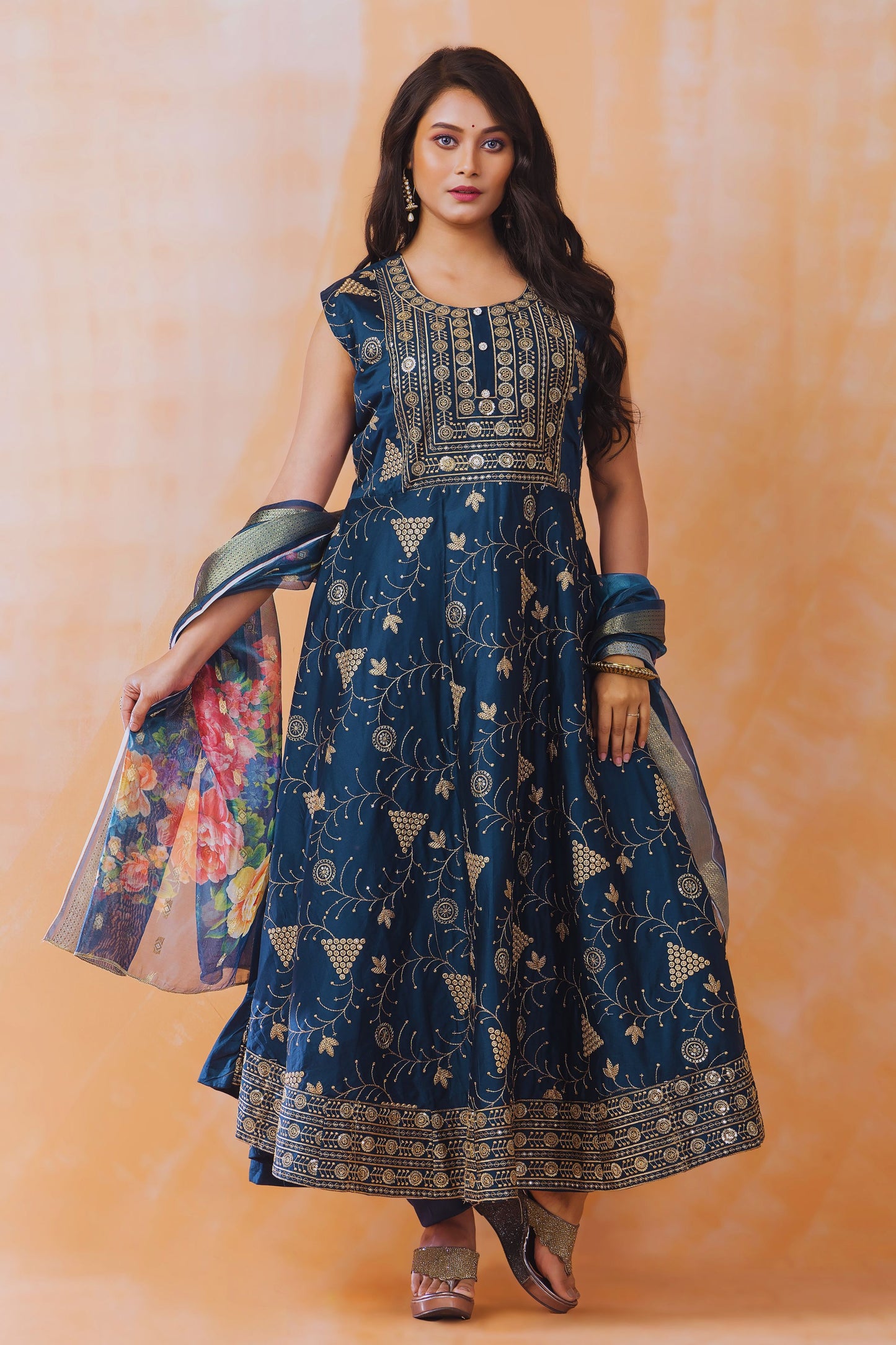 Woman wearing a blue embroidered traditional outfit with a floral stole against an orange background