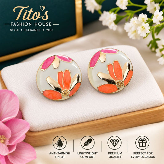 A pair of round gold-tone stud earrings with a white enamel background and a stylized orange and pink lotus flower design, displayed on a white cushion. The background includes a dark green gift box, white flowers, and a pink flower, with the brand logo 'Tito's FASHION HOUSE' in the top left and feature icons at the bottom.