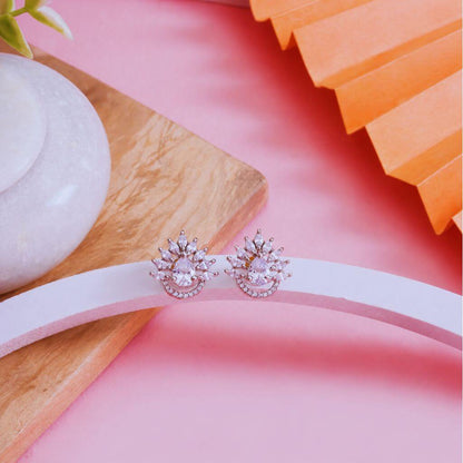 A pair of rose gold stud earrings with clear teardrop-shaped crystals and small clear accent stones, displayed on a white curved stand against a pink background with decorative props.