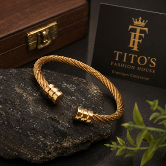 A gold-toned cable-wrapped cuff bracelet with two cylindrical end caps, displayed on a dark stone surface. A black card reading 'TITO'S FASHION HOUSE Premium Collection' and a wooden box are visible in the background, with small green leaves in the lower right corner.