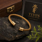 A gold-toned cable-wrapped cuff bracelet with two cylindrical end caps, displayed on a dark stone surface. A black card reading 'TITO'S FASHION HOUSE Premium Collection' and a wooden box are visible in the background, with small green leaves in the lower right corner.