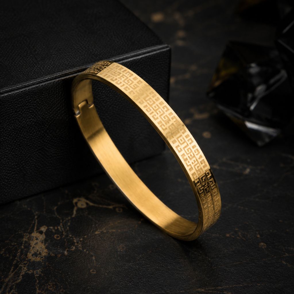A gold-plated metal bangle bracelet with engraved geometric patterns, resting on a dark textured surface next to a black box.