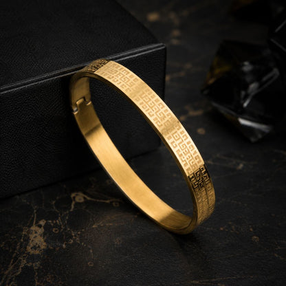 A gold-plated metal bangle bracelet with engraved geometric patterns, resting on a dark textured surface next to a black box.
