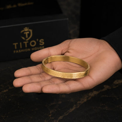 A gold-colored metal bangle bracelet with Greek key patterns on its outer surface, held in a person's hand. In the background, a black gift box with the brand name 'TITO'S FASHION HOUSE' is visible.