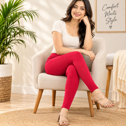 A woman sits in a light beige armchair wearing bright pink leggings, a white short-sleeve top, and beige heeled sandals in a bright, modern room with a potted plant and framed wall art.
