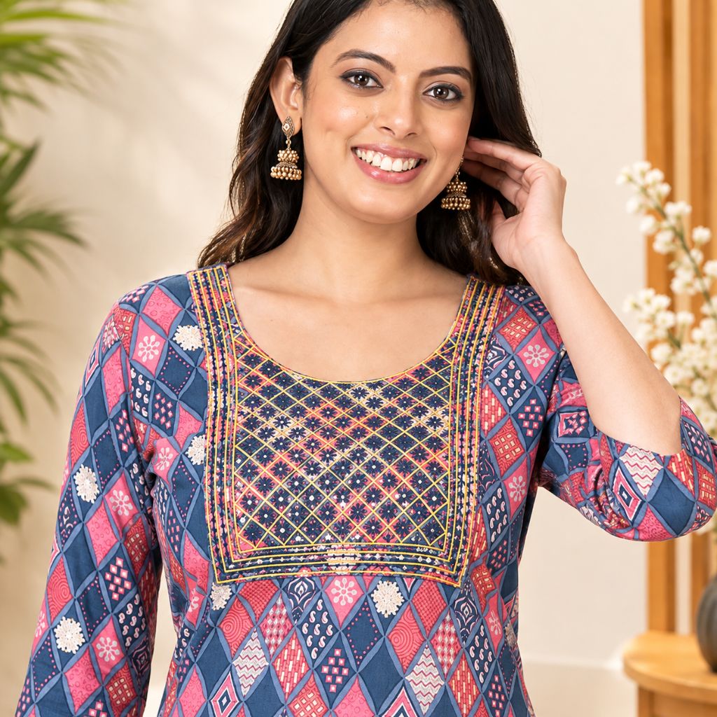 A woman wearing a navy blue kurta with red, pink, and gold geometric patterns and embroidery on the chest, paired with gold jhumka earrings, in a warm indoor setting.